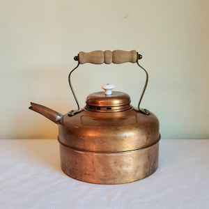 May include: A vintage copper kettle with a wooden handle and a white porcelain knob on the lid. The kettle has a tarnished finish and is in good condition.