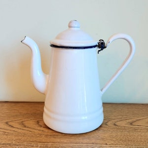 May include: White enamel coffee pot with a blue rim and a black handle. The pot has a rounded body and a spout on the side.