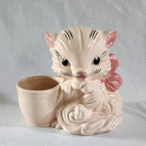 May include: A vintage pink ceramic cat planter with a small attached cup. The cat has black eyes, whiskers, and a pink bow. The planter is a light pink color and has a glossy finish. A charming decorative item.