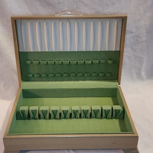 May include: Open wooden silverware storage box with a light tan exterior. The interior is lined with green felt and features slots for utensils. The top section has a light blue fabric backing.