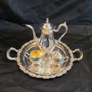 May include: A silver-plated tea service set consisting of a tall teapot, a creamer with a gold-toned interior, and a lidded sugar bowl. The pieces are arranged on a round, ornate silver tray featuring decorative handles and a detailed floral rim.