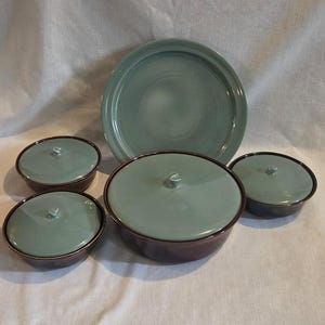 May include: A set of ceramic kitchenware featuring one large round platter, one large lidded bowl, and three smaller lidded bowls. Each piece has a glossy sage green interior and lid, contrasted by a dark brown exterior finish.