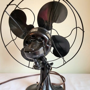 May include: An antique black electric fan with four blades, a protective cage, and a decorative base. The fan has a vintage design with a cord and plug.
