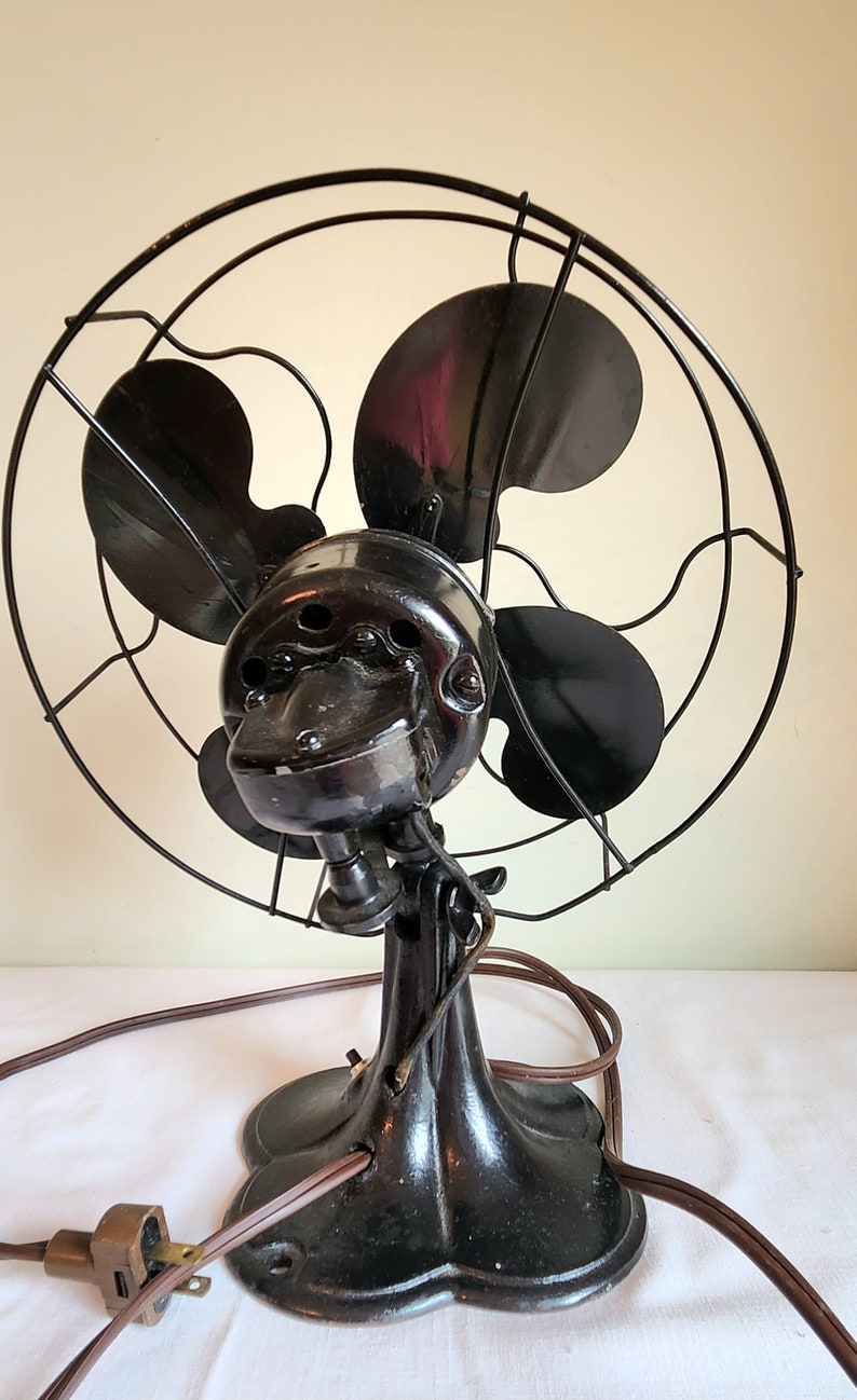 May include: An antique black electric fan with four blades, a protective cage, and a decorative base. The fan has a vintage design with a cord and plug.
