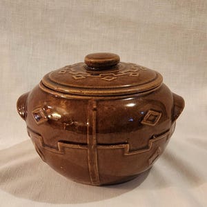 May include: A brown ceramic lidded pot with a round shape and two handles. The lid features a small knob and a geometric pattern. The pot's body has a repeating geometric design. The pot is likely for cooking or serving.