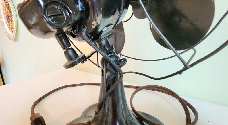 May include: An antique black electric fan with three blades and a protective wire cage. The fan has a dark brown power cord and a decorative base. The fan's motor housing shows signs of wear, adding to its vintage appeal.
