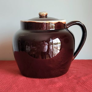 May include: A brown ceramic bean pot with a lid and a handle. The pot has a glossy finish and is decorated with a white rim.