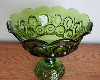May include: A green glass pedestal bowl with a scalloped rim and a starburst pattern. The bowl is on a wooden surface.