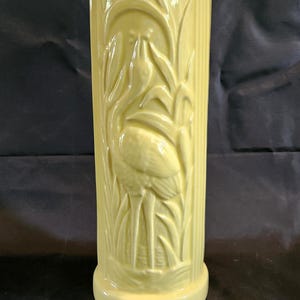May include: A tall, cylindrical, yellow ceramic vase with a flamingo and reeds design. The vase has a ribbed texture and a round base. The flamingo is depicted in a stylized, Art Deco manner.