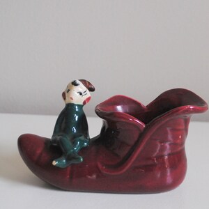 May include: A ceramic Christmas decoration featuring a burgundy sleigh with a seated elf figurine. The elf wears a green outfit and has a painted face. The sleigh has a textured surface and is designed for decorative purposes.