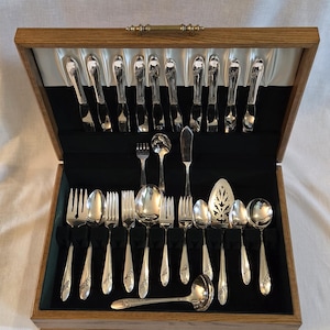 May include: A wooden silverware chest with a complete set of silver-colored flatware. The set includes forks, spoons, knives, and serving utensils, all neatly arranged on a black velvet-lined interior. The silverware has a decorative floral pattern.