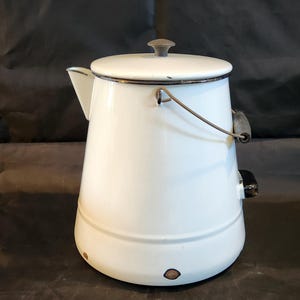 May include: A white enamel coffee pot with a lid and handle. The pot has a spout and a dark handle. The pot is white with a dark rim and a dark knob on the lid. The pot is likely vintage or antique.