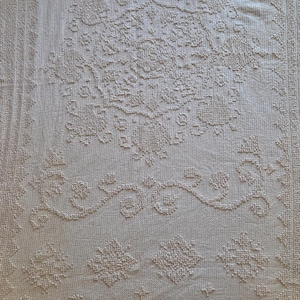 May include: A rectangular off-white textile featuring a raised, textured pattern. The design includes a central floral medallion, scrolling vine motifs, and geometric diamond shapes, all bordered by a repeating scalloped edge and linear bands.