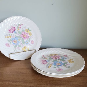 May include: Set of four white ceramic dinner plates with a floral design featuring pink, blue, and yellow flowers. The plates have a wavy edge.