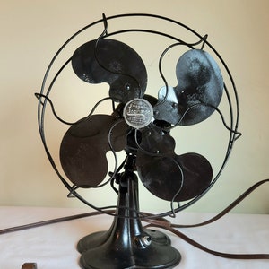 May include: An antique Emerson Seabreeze electric fan. The fan has four black blades and a black metal cage. The base is a dark, ornate design. The fan has a brown power cord and a two-prong plug. The fan's motor housing has a silver emblem.