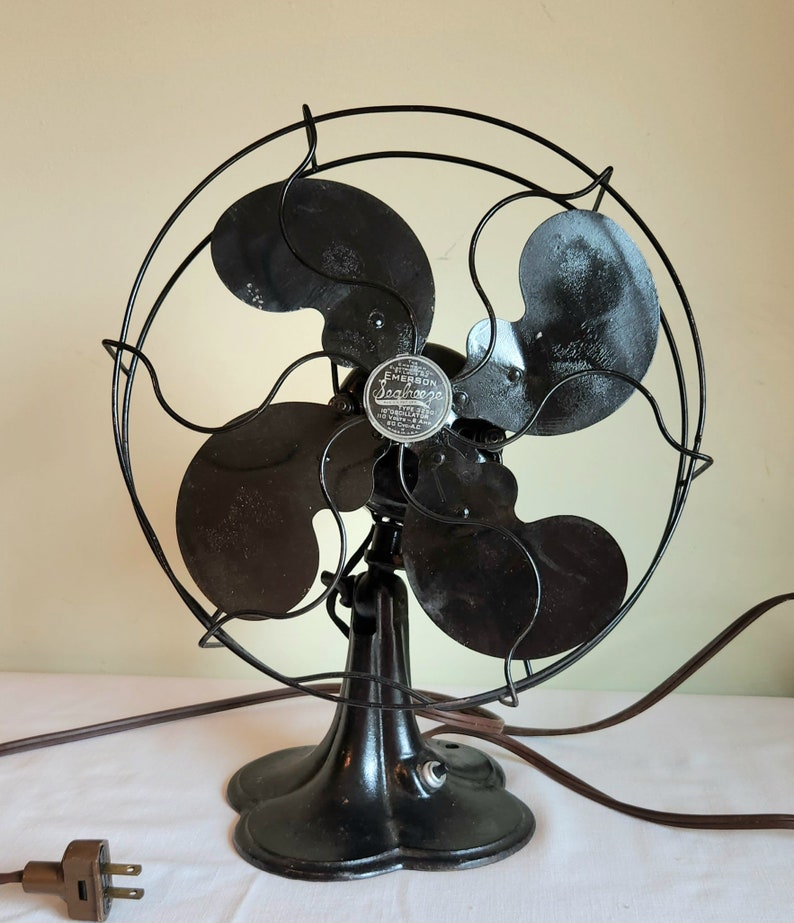 May include: An antique Emerson Seabreeze electric fan. The fan has four black blades and a black metal cage. The base is a dark, ornate design. The fan has a brown power cord and a two-prong plug. The fan's motor housing has a silver emblem.