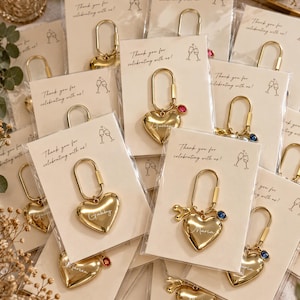 May include: A collection of gold-toned heart-shaped keychains attached to individual cards. Each card features the text 'Thank you for celebrating with us!' and a line drawing of two champagne glasses. Some keychains include small colored charms.
