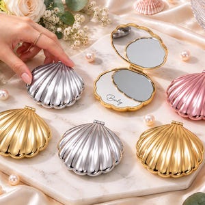 Pode incluir: Uma coleção de espelhos compactos em forma de concha com acabamentos metálicos dourados, prateados e rosa. Um espelho aberto exibe o nome Bailey em letra cursiva na superfície interna. Os compactos apresentam um design de borda festonada.