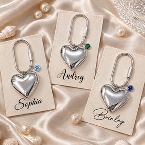 Può includere: Tre portachiavi a forma di cuore color argento con moschettoni ovali e piccoli ciondoli circolari con pietre portafortuna. Ogni portachiavi è montato su un cartoncino rettangolare beige con i nomi Sophia, Audrey e Brinley stampati in nero.