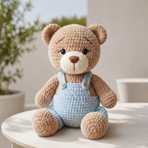 Può includere: Un orsacchiotto fatto a mano all'uncinetto, con corpo marrone chiaro e una tuta blu chiaro. L'orso ha un muso bianco, un naso marrone e occhi neri. L'orso è seduto su una superficie bianca.