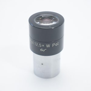 May include: A microscope eyepiece with a black upper section and a silver-colored lower section. The text "pl 12.5x W Pol" is printed on the black section. The eyepiece has a lens at the top.