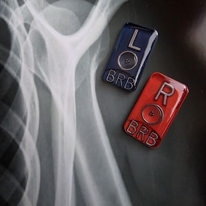 May include: Two rectangular enamel pins featuring silver lettering and circular accents. The blue pin displays the letter L and the text BRB, while the red pin displays the letter R and the text BRB. Both pins are positioned over a grayscale X-ray image.