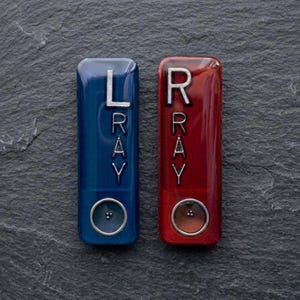 May include: Two rectangular hearing aid accessories, one blue and one red, each featuring the silver letters LRAY or RRAY vertically aligned. Each device includes a circular button at the bottom with three small dots in the center of the circle.
