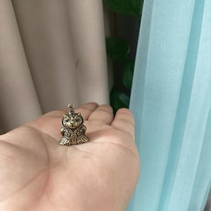 May include: A small, antique brass-coloured cat figurine with intricate details. The cat is wearing a decorative outfit and a crown. The figurine has a loop at the top for hanging. The cat is held in a person's hand.
