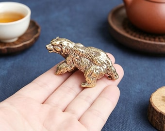 May include: A small, detailed brass bear figurine. The bear is depicted in a walking pose, with textured fur and a realistic appearance. The figurine is held in a person's hand, showcasing its size and craftsmanship. The bear is a decorative item.