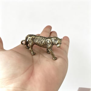 Op de afbeelding: Een kleine bronzen tijgerfiguur met gedetailleerde geëtste markeringen. De tijger wordt in een loopstand getoond. De figuur heeft een verweerde afwerking, waardoor hij een antieke uitstraling heeft. De tijger is een symbool van kracht en moed. Ongeveer 7 cm lang.