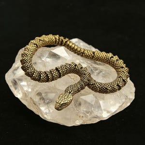 Puede incluir: Una figura de serpiente articulada en tono latón con una textura de escamas detallada en todo el cuerpo. La serpiente está enroscada en una posición curva, con una cabeza definida con ojos y líneas de boca visibles, sobre un cristal de cuarzo claro.