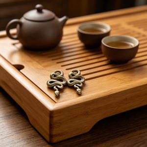 May include: A wooden tea tray with a brown teapot and two small cups. Two brass snake-shaped earrings are on the tray. The tea set is arranged on a wooden surface, suggesting a tea ceremony.