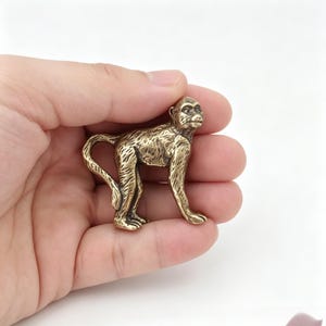 Könnte beinhalten: Kleine, detaillierte Affenfigur aus Messing. Der Affe ist in einer stehenden Pose mit eingerolltem Schwanz dargestellt. Die Figur hat eine strukturierte Oberfläche und eine warme, goldene Farbe. Es ist ein dekorativer Artikel.
