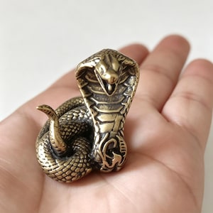 Könnte beinhalten: Detaillierte Cobra-Figur aus Messing, zusammengerollt und bereit zum Zuschlagen. Die Haube der Schlange ist erhoben und ihre Schuppen sind aufwendig detailliert. Die Skulptur hat ein Vintage-Aussehen mit einer satten, goldbraunen Farbe. Die Kobra ist ein Symbol für Macht und Schutz.