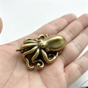 May include: A detailed brass octopus figurine with eight curled tentacles and a rounded head. The octopus is a golden color and appears to be made of metal. The figurine is resting on a palm.