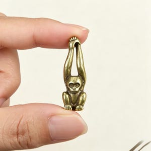Puede incluir: Una pequeña figura de mono de metal color latón representada en posición colgante. El mono tiene los brazos extendidos hacia arriba, formando un bucle en la parte superior, con el cuerpo y la cara claramente definidos en un acabado metálico dorado.