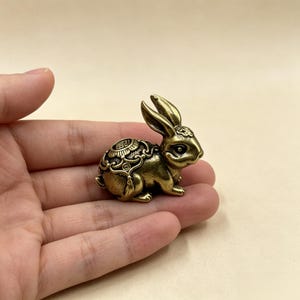 May include: A small, antique-style brass rabbit figurine featuring intricate embossed patterns on its back and head. The rabbit is depicted in a seated position with long, upright ears and detailed facial features, showcasing a metallic gold finish.