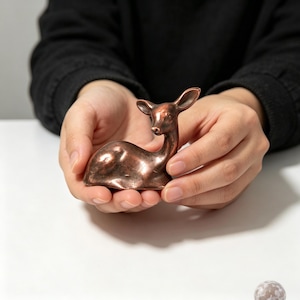 Puede incluir: Una pequeña figura metálica de color cobre que representa a un ciervo en posición de reposo. El ciervo tiene contornos suaves y curvos, orejas puntiagudas y una expresión facial suave, capturando al animal en una pose tranquila y estacionaria.