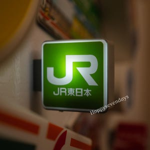 Peut inclure: Une enseigne carrée lumineuse avec un fond vert vif, arborant le logo blanc JR et le texte japonais JR東日本 en dessous. L'enseigne possède un cadre en argent métallique et est fixée sur un support sombre, diffusant une lueur douce.