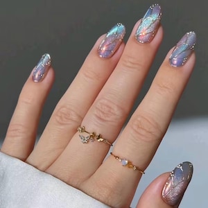 Peut inclure: Gros plan sur une main ornée de longs ongles ovales. Les ongles présentent un motif chatoyant aux tons bleus, violets et dorés, évoquant des ailes de papillon. Deux bagues en or, l'une avec un papillon, l'autre avec une petite pierre précieuse.