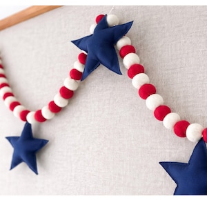 May include: A patriotic garland featuring alternating red and white felt balls, interspersed with navy blue star-shaped ornaments. The garland is displayed against a neutral background, perfect for festive decor.