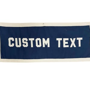 May include: A rectangular navy blue banner featuring the white text CUSTOM TEXT centered in a bold, block font. The banner is framed by a thick, off-white canvas border and includes three metal grommets along the top edge for hanging purposes.