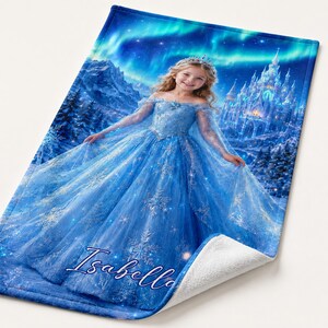Op de afbeelding: Een blauwe deken met een meisje in een blauwe jurk en kroon, tegen een achtergrond van een kasteel en het noorderlicht. De naam "Isabella" staat in cursief op de deken.