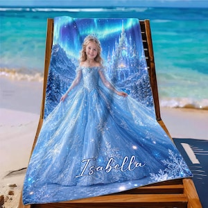 Op de afbeelding: Een blauwe strandlaken met een jong meisje in een blauwe jurk met een kasteel en een aurora borealis achtergrond. De handdoek heeft de naam "Isabella" in witte letters. De handdoek ligt op een houten strandstoel.