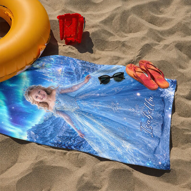 Op de afbeelding: Een strandlaken met een blauw en wit ontwerp, met een jong meisje in een blauwe jurk en de naam "Isabella". De handdoek is versierd met sneeuwvlokken en een sterrenhemel. Zonnebril en oranje slippers liggen op het zand.