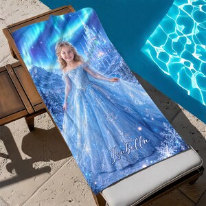 Op de afbeelding: Een blauwe strandlaken met een afbeelding van een meisje in een blauwe jurk, een kasteel en het noorderlicht. De naam "Isabella" staat op de handdoek. De handdoek ligt op een houten ligstoel naast een zwembad.