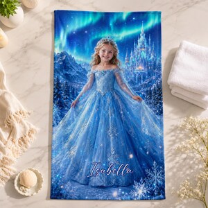 Op de afbeelding: Een blauwe badhanddoek met een afbeelding van een meisje in een blauwe jurk, een tiara en een kasteel. De handdoek heeft een winterthema met sneeuwvlokken en een aurora borealis-ontwerp. De naam "Isabella" staat op de handdoek.