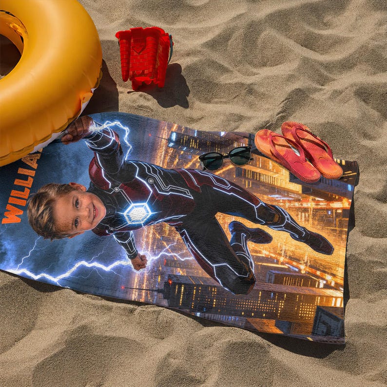 Puede incluir: Una toalla de playa con un dise&ntilde;o de superh&eacute;roe, que presenta la imagen de un ni&ntilde;o, rayos y un horizonte urbano. La toalla est&aacute; sobre arena, con gafas de sol y chanclas. Tambi&eacute;n hay un flotador amarillo y un cubo rojo.