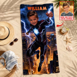 Puede incluir: Una toalla de playa personalizada con un dise&ntilde;o de superh&eacute;roe. Presenta a un ni&ntilde;o con un disfraz con rayos y un fondo de ciudad. El nombre "WILLIAM" est&aacute; impreso en la parte superior. Una peque&ntilde;a foto del ni&ntilde;o est&aacute; en la esquina. La toalla est&aacute; en una playa de arena.