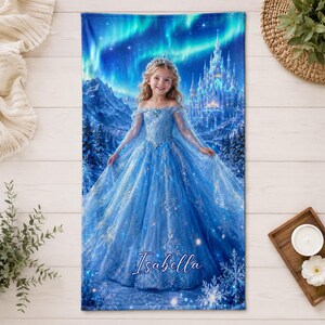 Op de afbeelding: Blauw strandlaken met een jong meisje in een blauwe jurk, tiara en cape, tegen een kasteel en een winterlandschap. De naam "Isabella" is cursief geschreven. De achtergrond toont het noorderlicht.